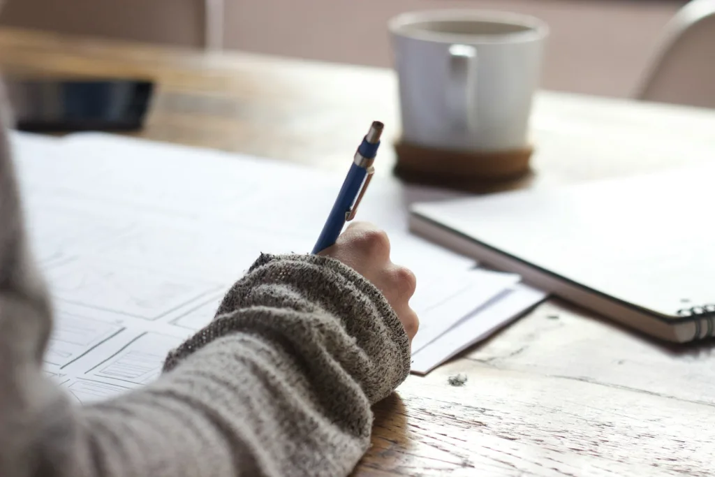 person writing on brown wooden table near white ceramic mug